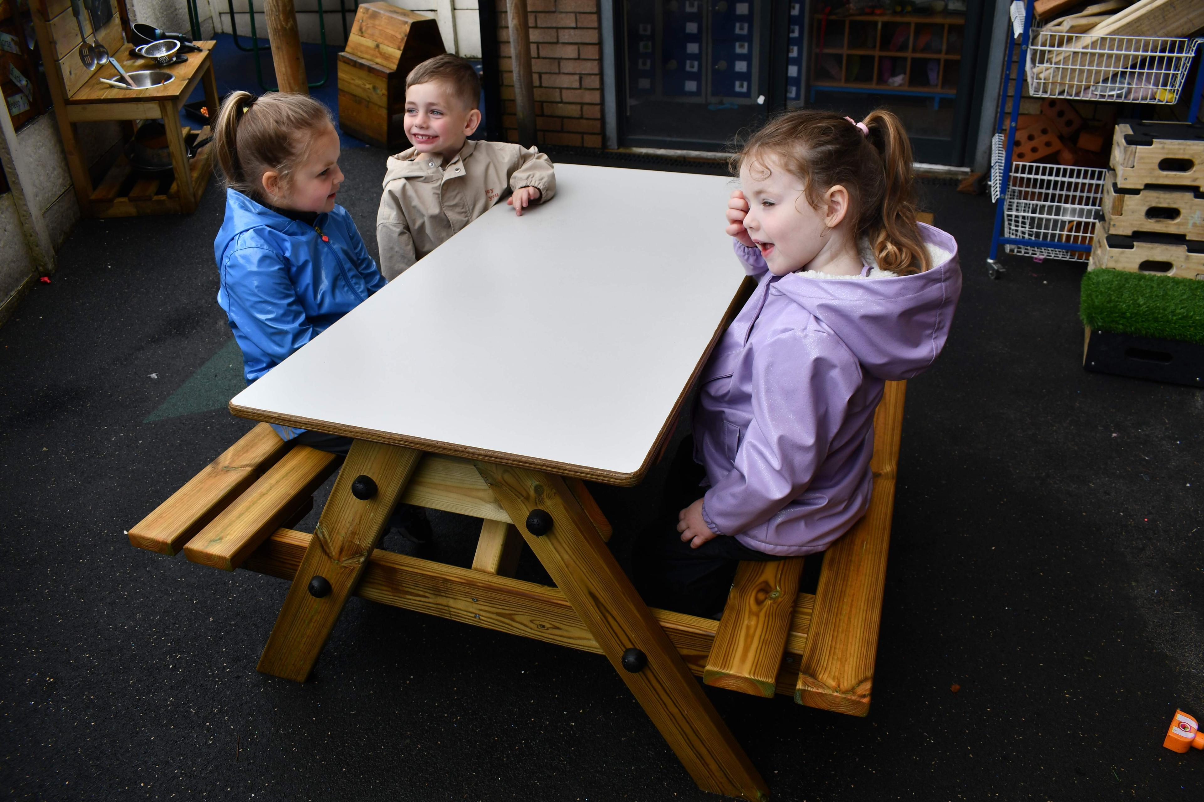3 children are sat at the EYFS Picnic Table. A girl and boy are sat on the left, smiling and looking around whilst the girl on the right side is talking to someone off camera.
