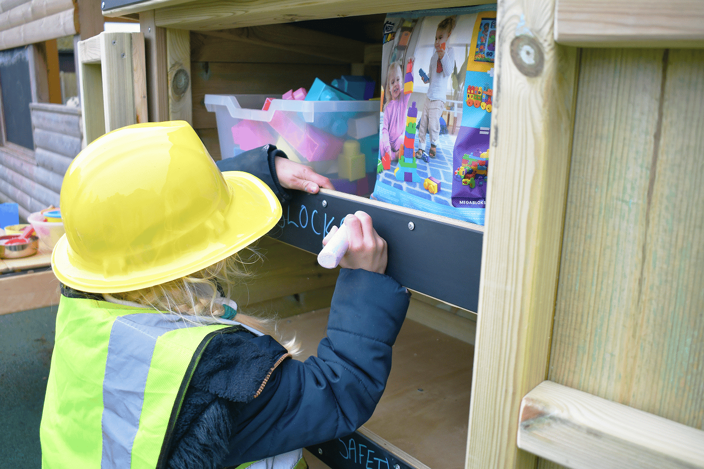 Young school writing with chalk on the Wooden Acorn Storage Unit in a school playground