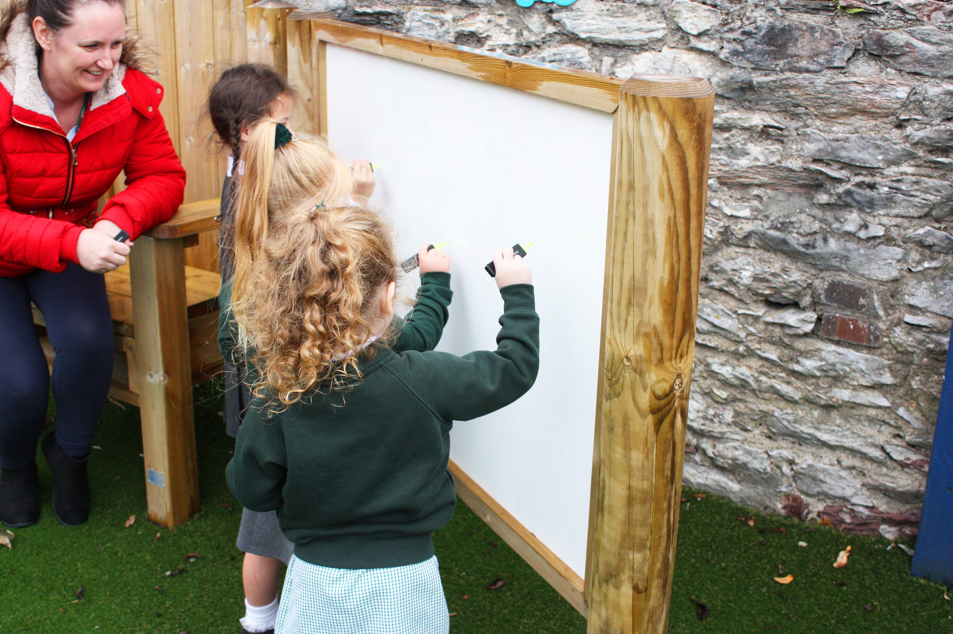 3 children are holding highlighters and are going to draw on a big whiteboard. The whiteboard has a wooden border around it, with a teacher sat on a big wooden chair next to it.