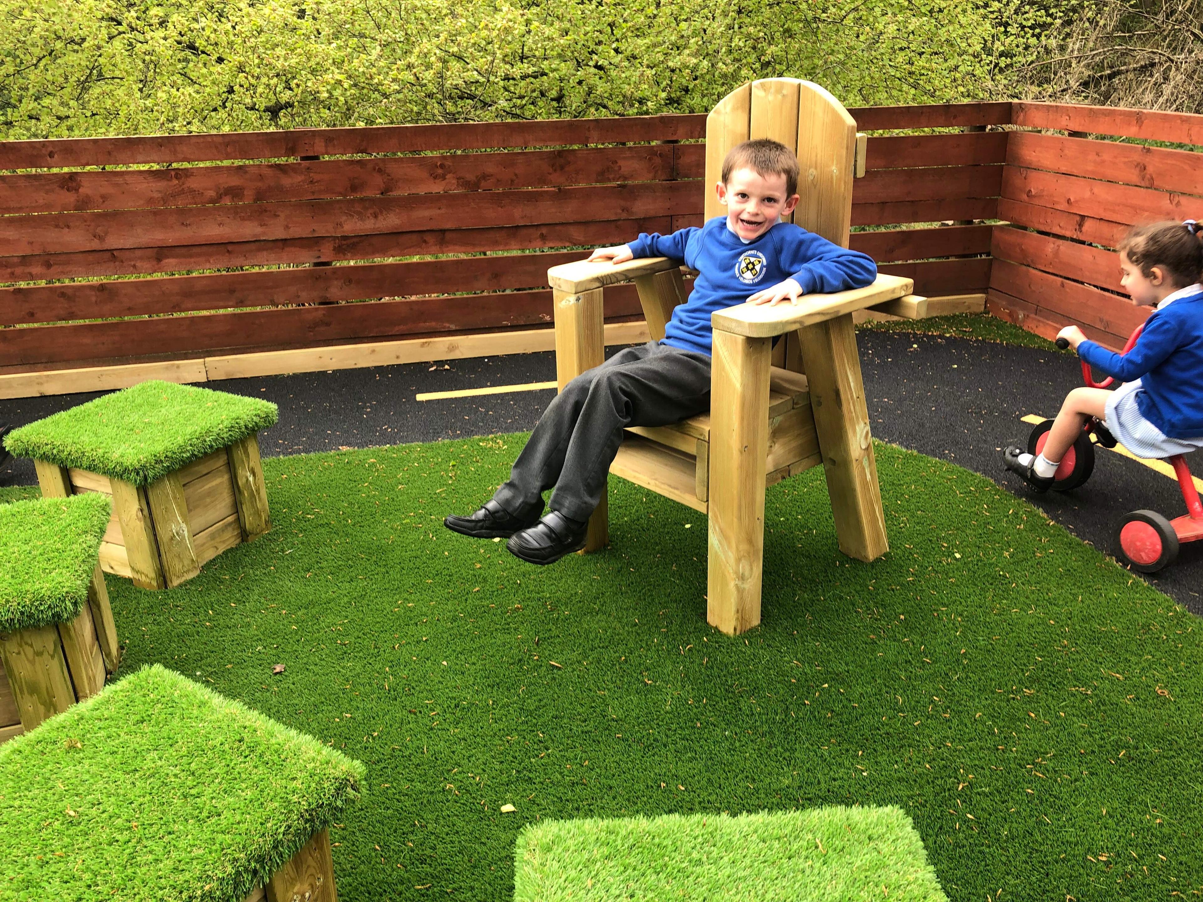A child is sat in a big wooden chair, opposite 4 wooden blocks with artificial grass on the top of them. The chair and blocks are placed on an artificial grass surface.