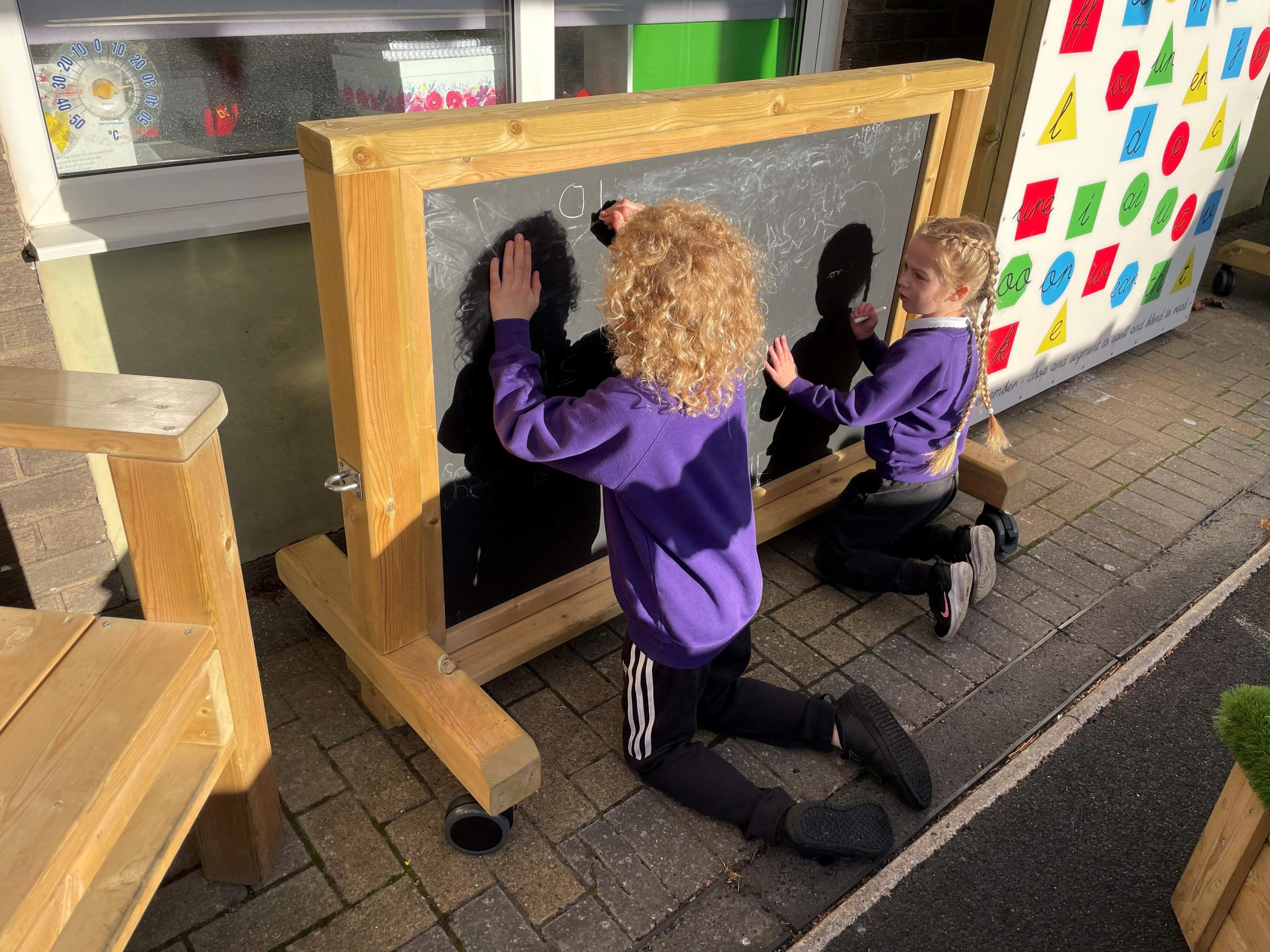 2 children are kneeling as they hold chalk. They are facing a big chalkboard. One child has drawn the numbers 0 and 1. The other child is watching, ready to start drawing too.