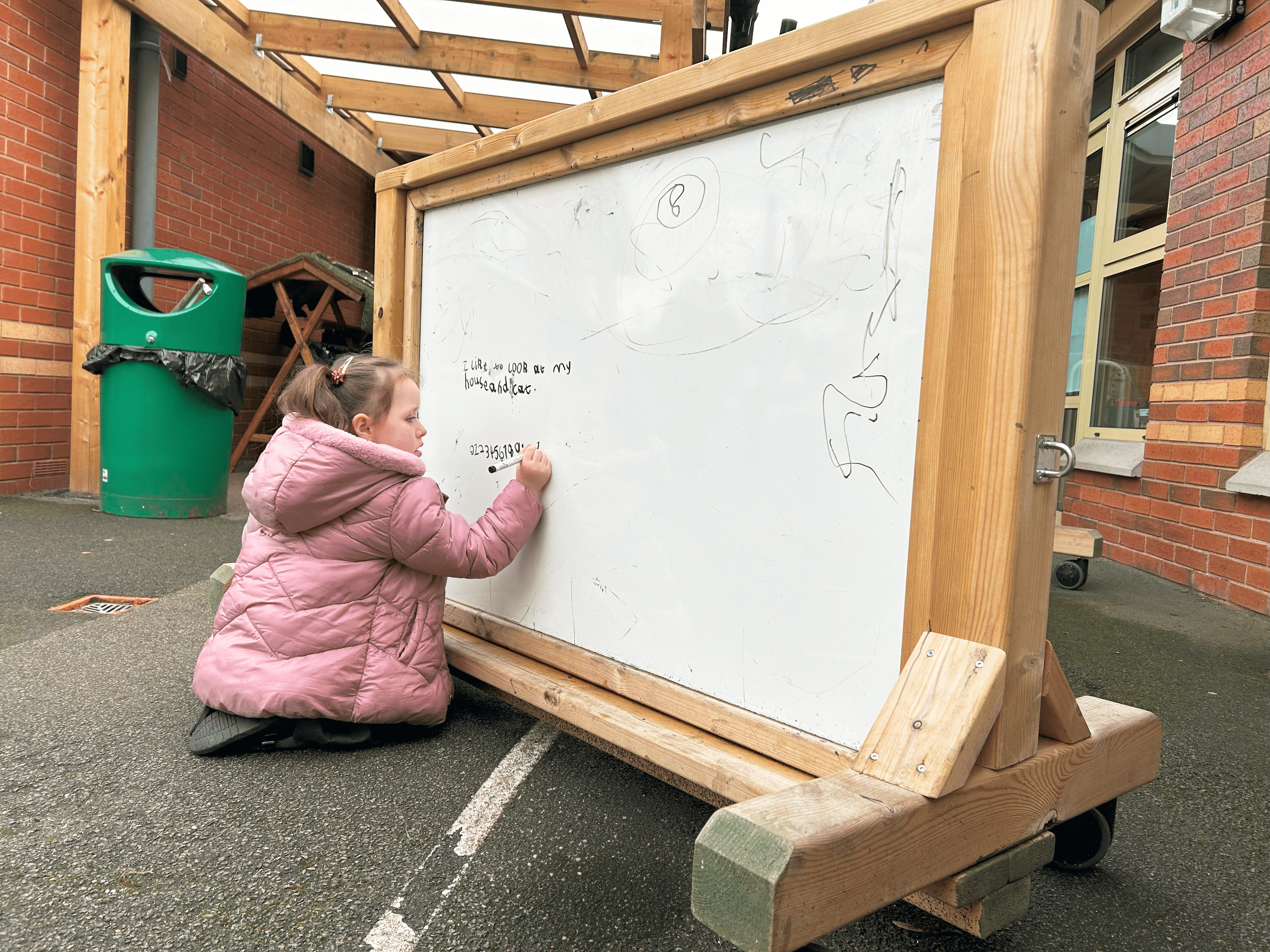 A little girl is on her knees as she holds a whiteboard marker. She is opposite as whiteboard as she is writing on the board. She's written "I like to look at my house and cat" and wrote numbers.