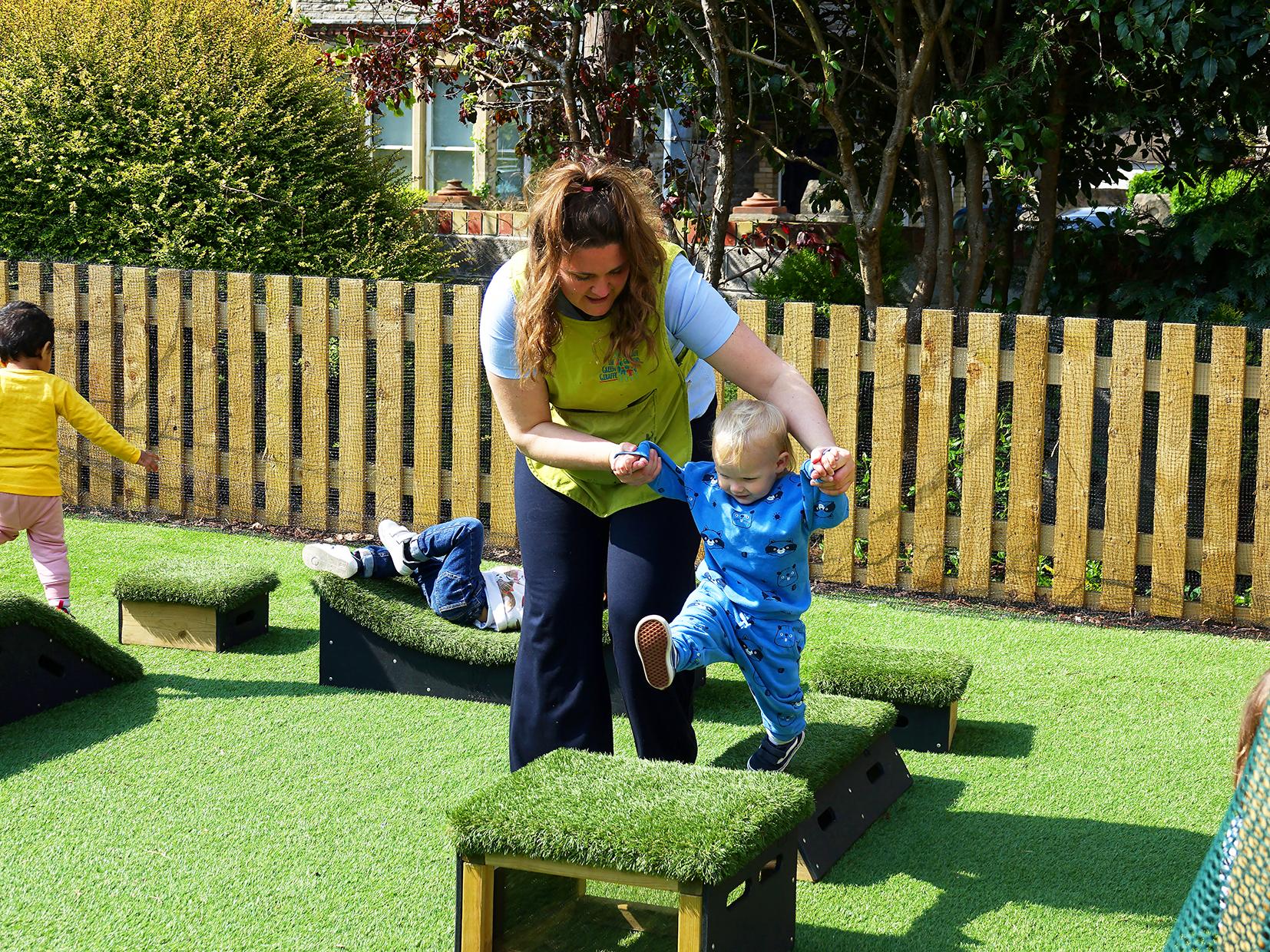 A toddler is being helped by an adult to play with the Get Set, Go! Blocks. The child's hands are being held as they are supported as the toddler steps to the next block.