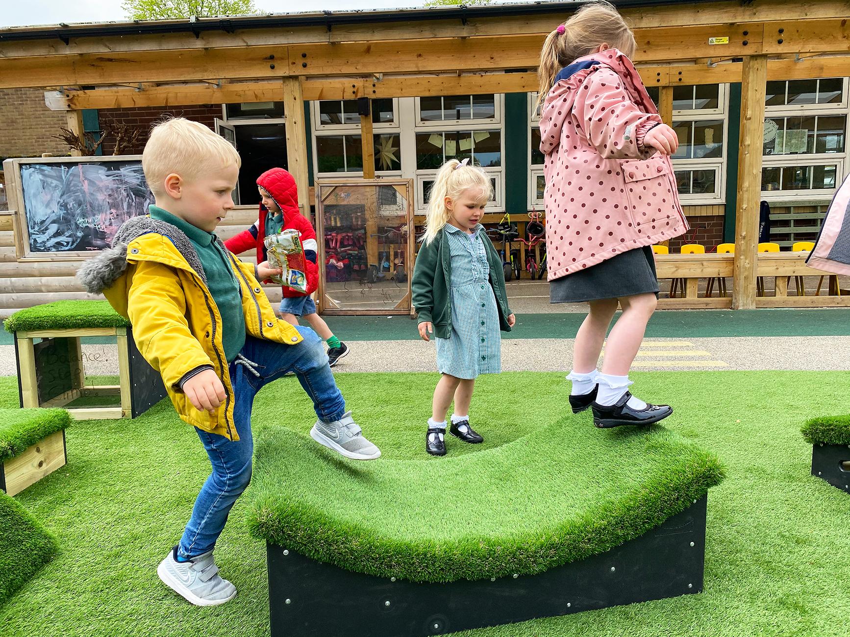 4 Children are stood around one of the Get Set, Go! Blocks, which is shaped like a half-pipe skate ramp. Two children are stepping on to the block, whilst the other two children are looking around.