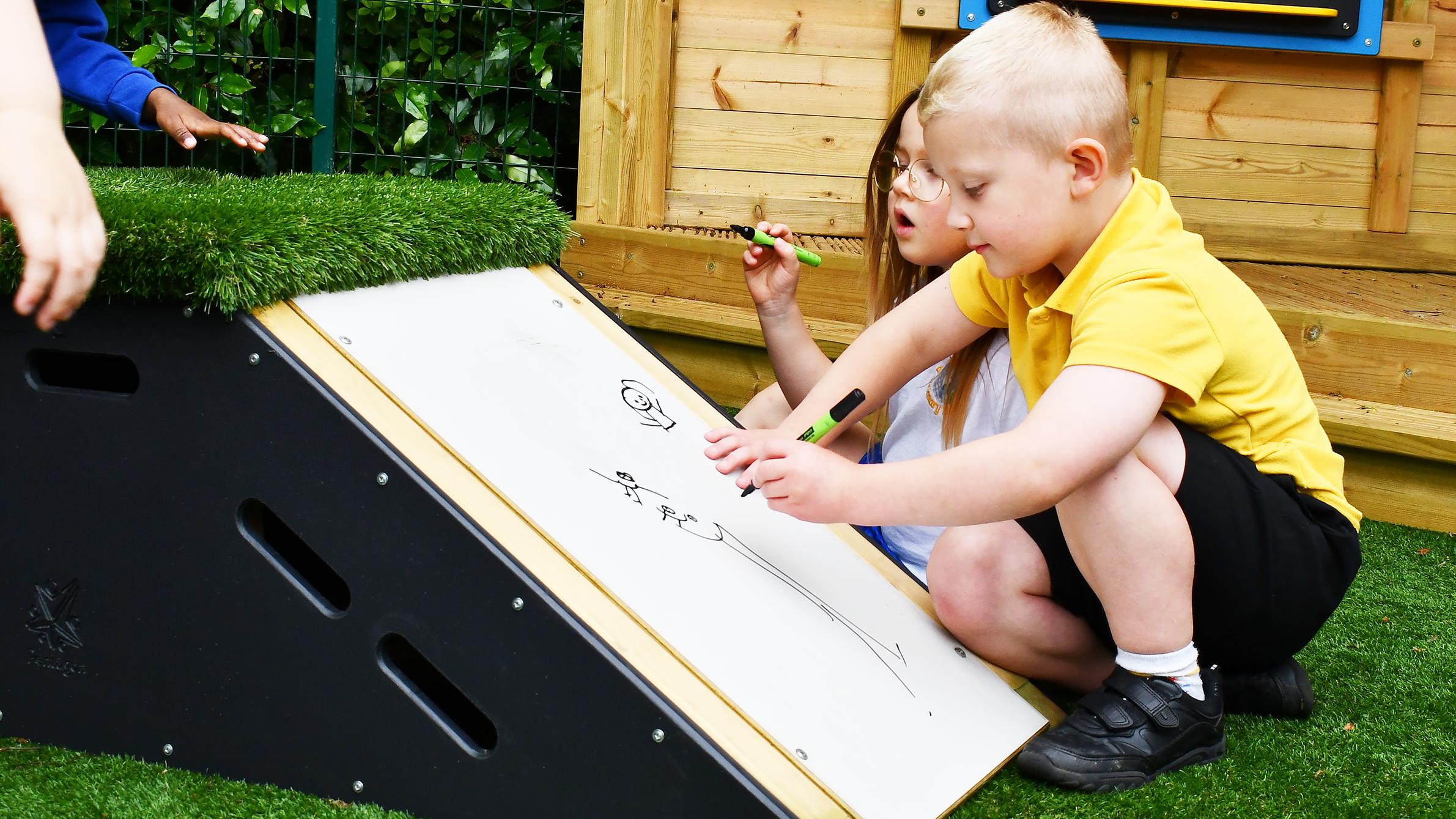 2 children are kneeling as they draw on a PTEG mark making panel that is placed on a ramped block.