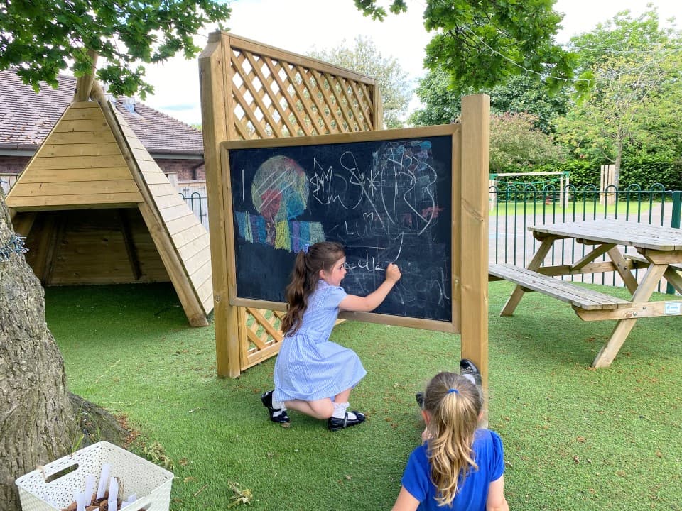 two children drawing on the giant chalkboard with chalk as they sit on the artificial grass 