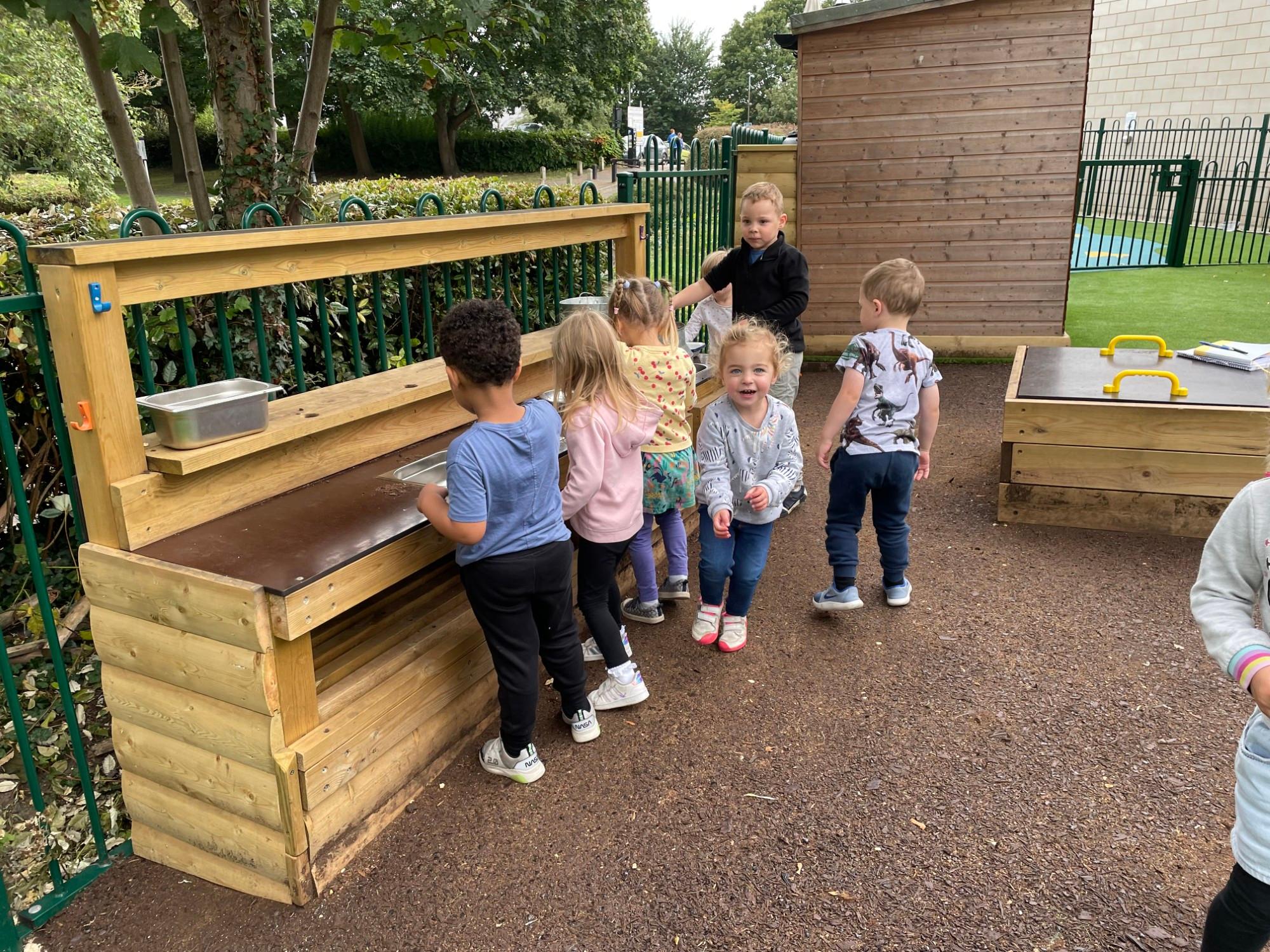 6 nursery children gathered around a mud kitchen