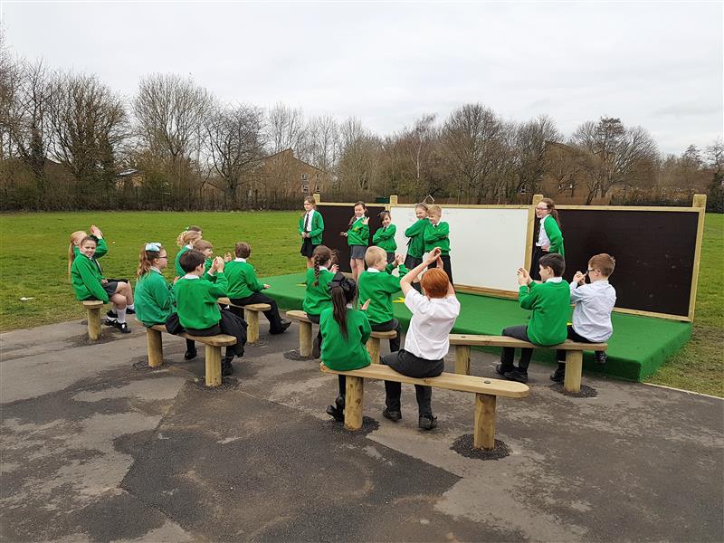 6 children are on an outdoor performing arts stage at school. 14 children are sat on 7 wooden benches, which have all been placed at the front of the stage. The audience are all clapping as the 6 children are posing.