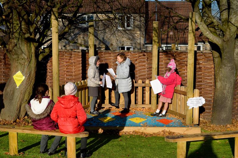 3 girls are on an outdoor performance stage, built by Pentagon Play. They are holding scripts and are performing in front of 2 other children, who are sat on a bench opposite the stage.