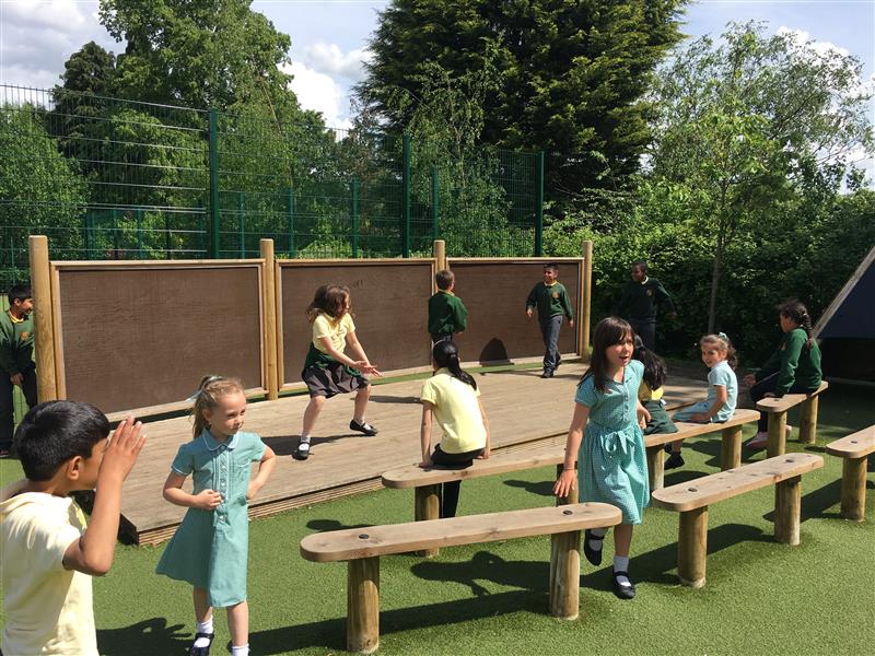 3 children are playing on the outdoor school stage, which was installed by Pentagon Play. 8 children are stood around the front of this performance and are watching and talking to each other,