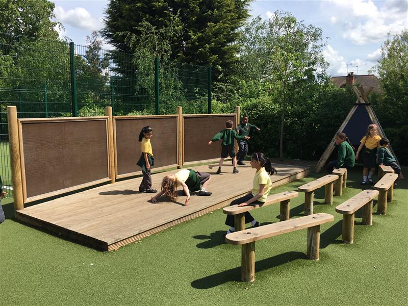 4 children are performing on an outdoor wooden stage. As the 4 children are performing, 4 other children can be seen sat on wooden benches, Infront of the stage. They are watching the play.