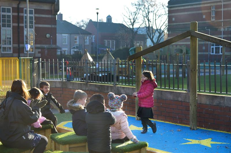 One child is on the outdoor stage, which was made by Pentagon Play, and is doing a performance. There are 5 children and 1 teacher in the audience. They are all sat on wooden benches and are clapping to the child performing.