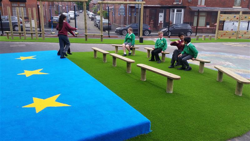 2 children are running off a blue outdoor stage, with star designs on it. 4 children are sat on wooden benches and are watching the play that is happening in front of them.