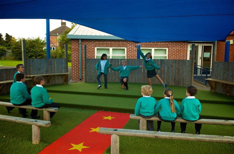 Three children are on an outdoor stage, which is covered by a blue canopy and seem to be performing a dance. There are 6 children who are sat on benches in front of the stage and are watching the performance.