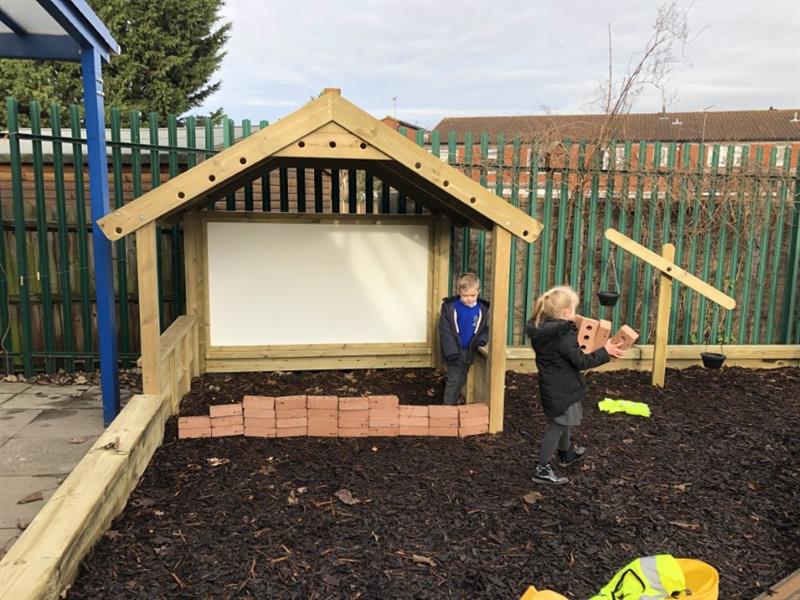 One boy stood inside a giant playhouse with a whiteboard, behind a small wall of bricks wearing a blue jumper and black coat whilst one girl wearing a black coat carries 3 bricks across the dark brown bark. 