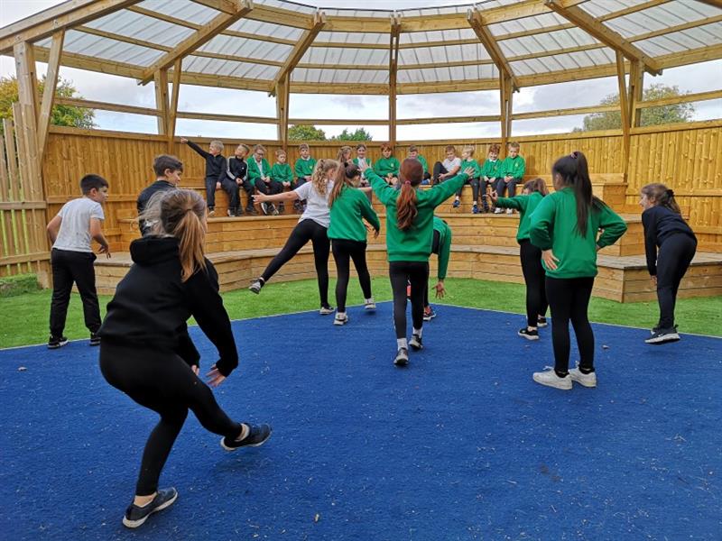 Children performing on a performance stage under a huge amphitheatre with more children sat on the seats in the audience. 