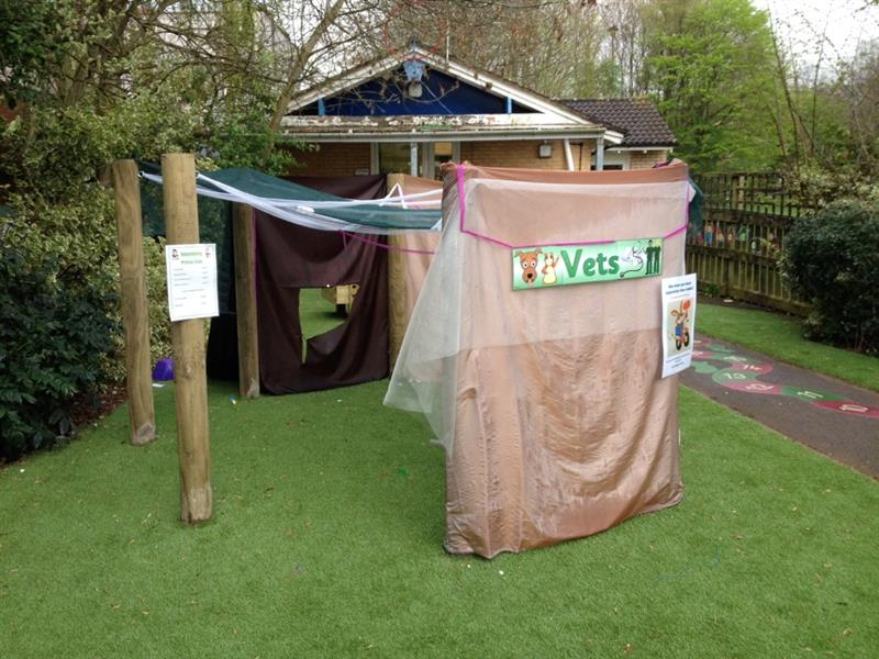 A creative den made using den making poles, a dark brown sheet and a light brown sheet with a poster stuck on saying 'vets'. Den has been made next to a snake playground marking with numbers on it. 