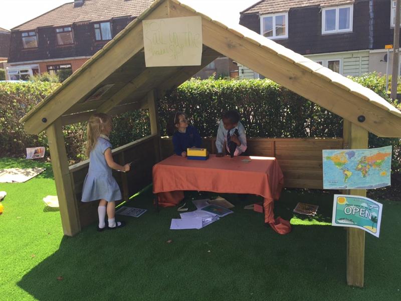 3 children stood behind a small table with a red cloth on top, inside a giant playhouse which they have made into a little shop. The playhouse has been installed on artificial grass. 