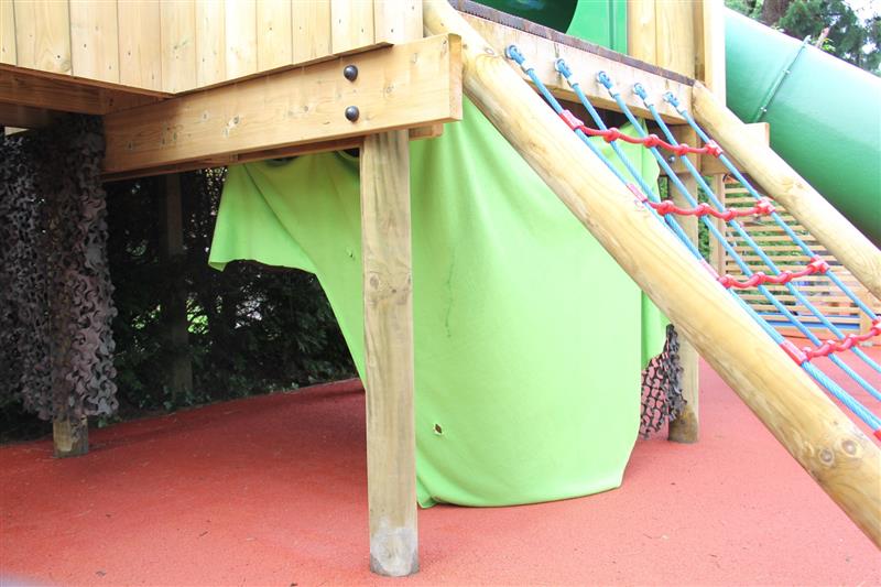 A creative den made using a lime green sheet and camouflage netting underneath a large timber play frame which has been installed onto red surfacing with a green slide attached to the play frame. 