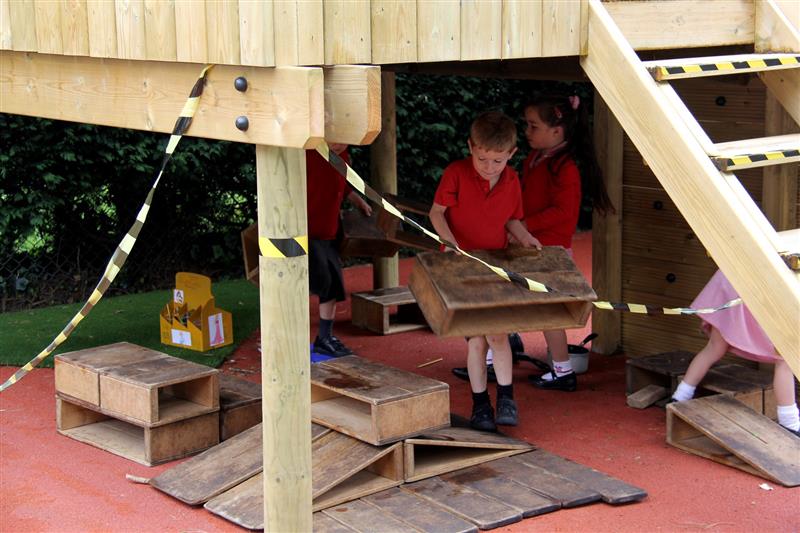 4 children, 3 wearing red tops and one girl wearing a red summer dress playing underneath a large timber play frame with one boy carrying a wooden box on red playground surfacing. 