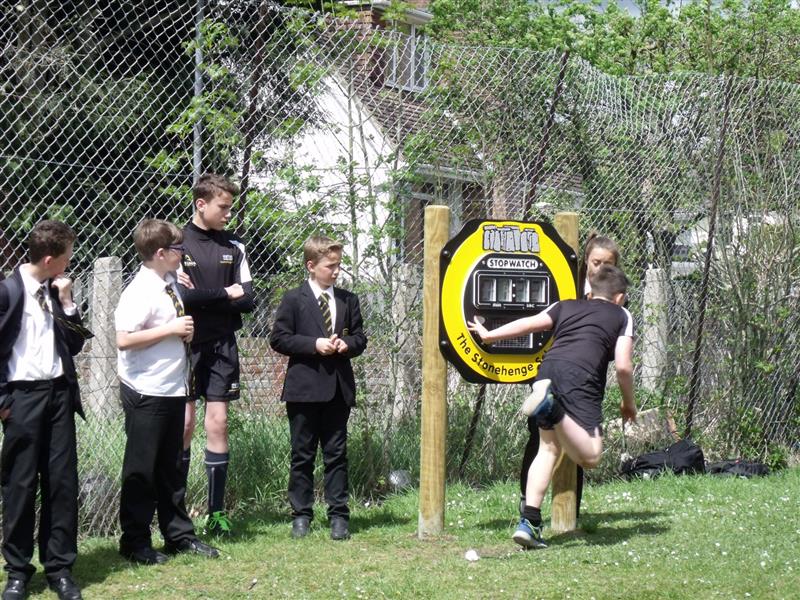 5 children wearing school uniform stood next to a large stopwatch on timber posts installed into grass whilst one boy runs past touching the stop watch.