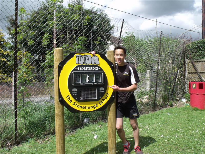 One boy smiling and standing infront of large stopwatch on timber posts which has been installed into grass, he is wearing a PE kit with black trainers and red shoe laces. 