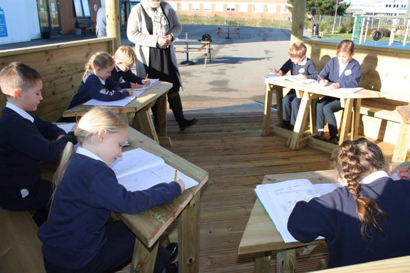 Children sat at work stations in a timber outdoor gazebo working in their work books