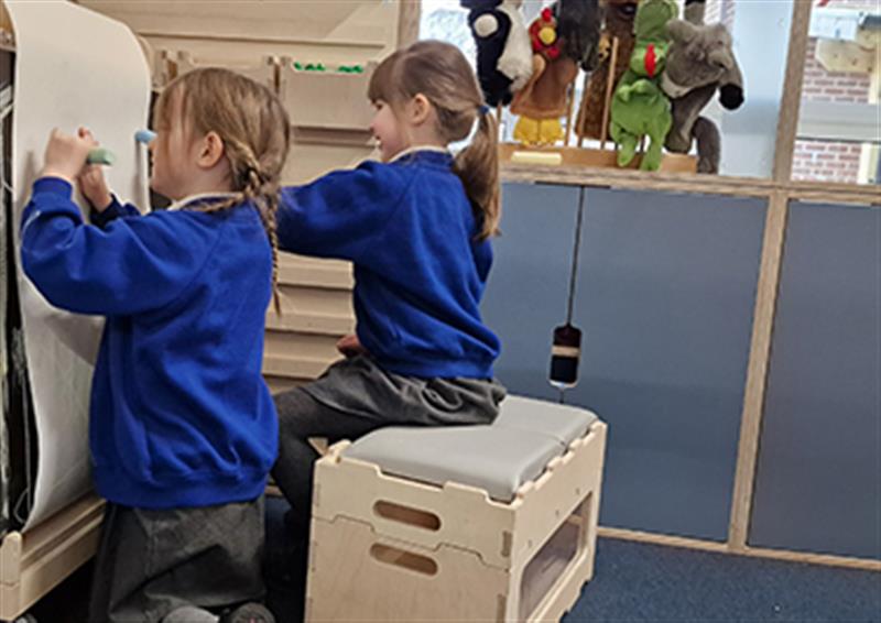 Two young girls sat on a Stack and Sit bench drawing on the Art Easel paper in a classroom 