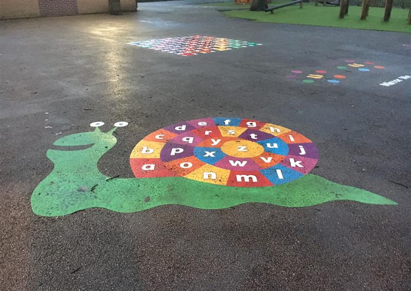A thermoplastic playground marking of a big snail, with the shell showing the alphabet in a spiral motion. Each letter has a different colour background, with the snail itself being a green colour, with eyes and a smile. The graphic is placed on a tarmac playground, with other playgrounds around.