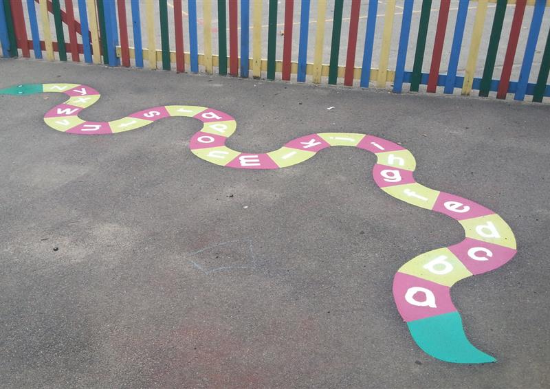 A thermoplastic snake marking has been installed on a tarmac playground. The snake has a green head and tail, but it's body is broken in to blocks that alternate between yellow and red. Each block contains a letter in alphabetical order, with a being at the tail.