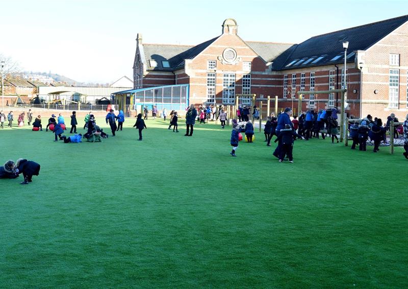 A field of artificial grass with hundreds of children playing on it. The children are walking and playing all over the area, with little groups forming. Buildings can be seen in the background.