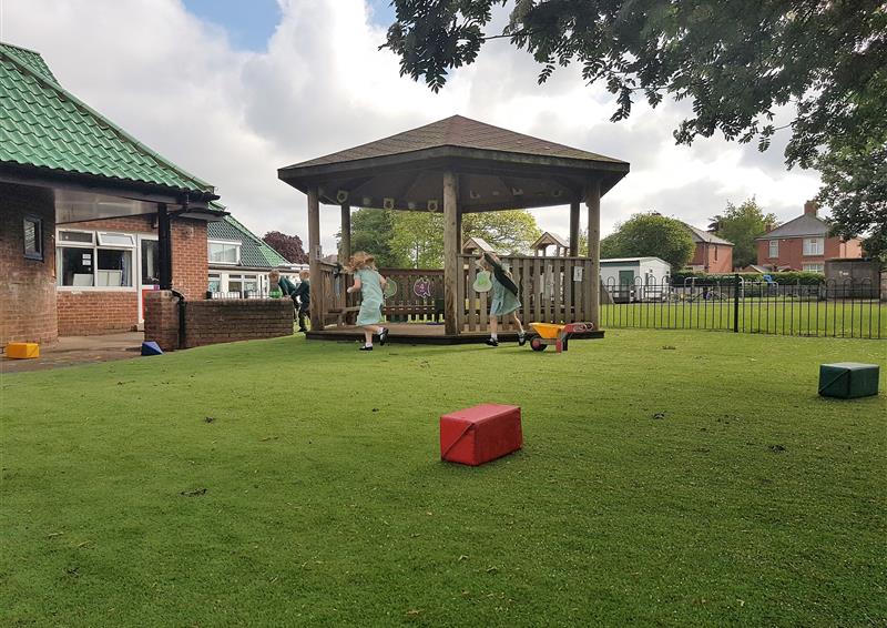 Artificial grass area with a wooden canopy in the centre. A group of children are playing around the canopy. The school can be seen to the left of the artificial grass area.