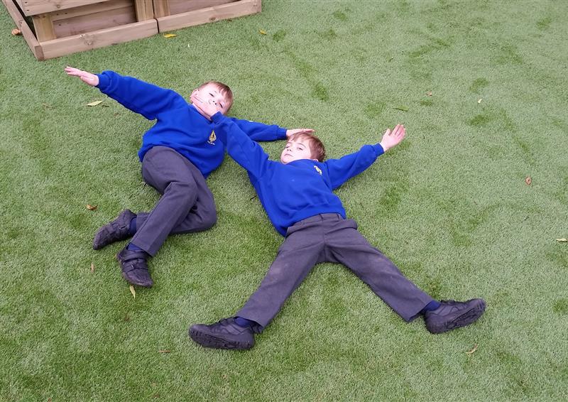 A surface of artificial grass with two children lying down in a star pose. The children are looking at the camera and smiling.