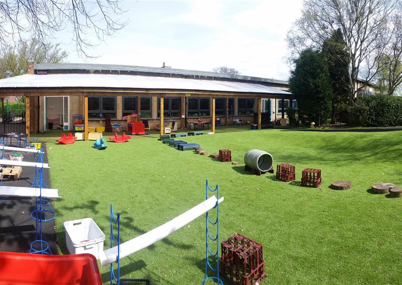 A field of artificial grass with a variety of objects placed in a line. These objects form an obstacle course for children to play on. The school can be seen in the background.