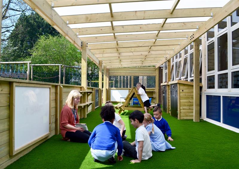 A group of children are sat on artificial grass and are listening to a teacher. A timber canopy covers the top of them, providing them shade from the sun. A variety of outdoor play equipment can be seen in the background, with a whiteboard present next to the teacher.