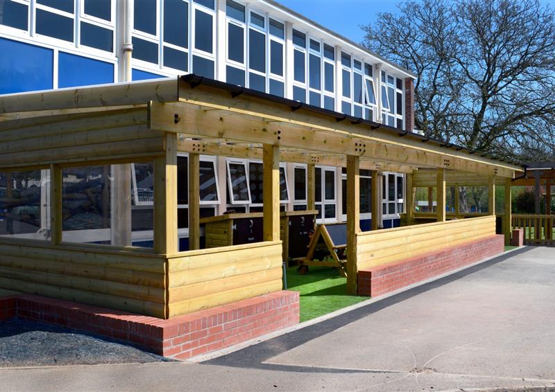 A playground timber canopy that has been installed on a concrete playground. The inside surfacing is artificial grass and has a variety of outdoor learning equipment inside. It is also connected to the main school building.
