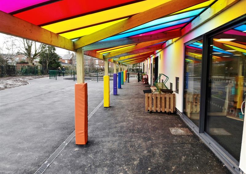A timber canopy is attached to the side of a school building, The roof of the timber canopy is made of a variety of different coloured plastic sheets, creating a sensory zone.