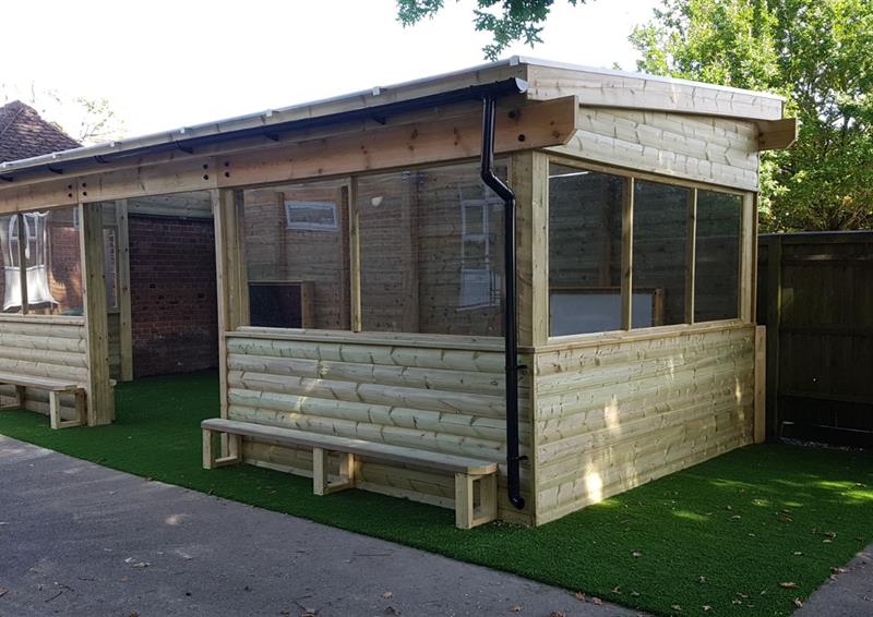 A timber outdoor canopy, which is freestanding. The canopy has walls and windows, creating it's own building. The flooring is artificial grass, with the outside surface being tarmac. The canopy is surrounded by nature.