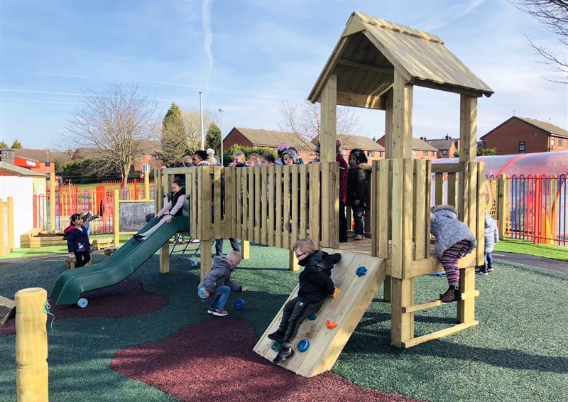 A play tower that has been installed on a wetpour surface. A red path can be seen coming from the climbing wall and slide, with the rest being green wetpour. A big group of young children are playing on the equipment. The school can be seen in the background.