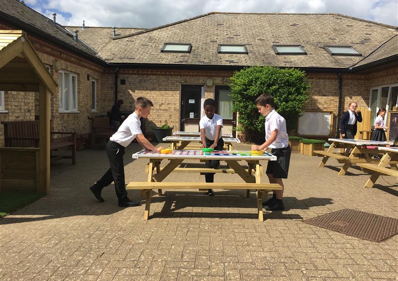 Three children are stood around a picnic table with a gametop. They are playing a board game that has been printed into the tabletop and the school can be seen in the background.