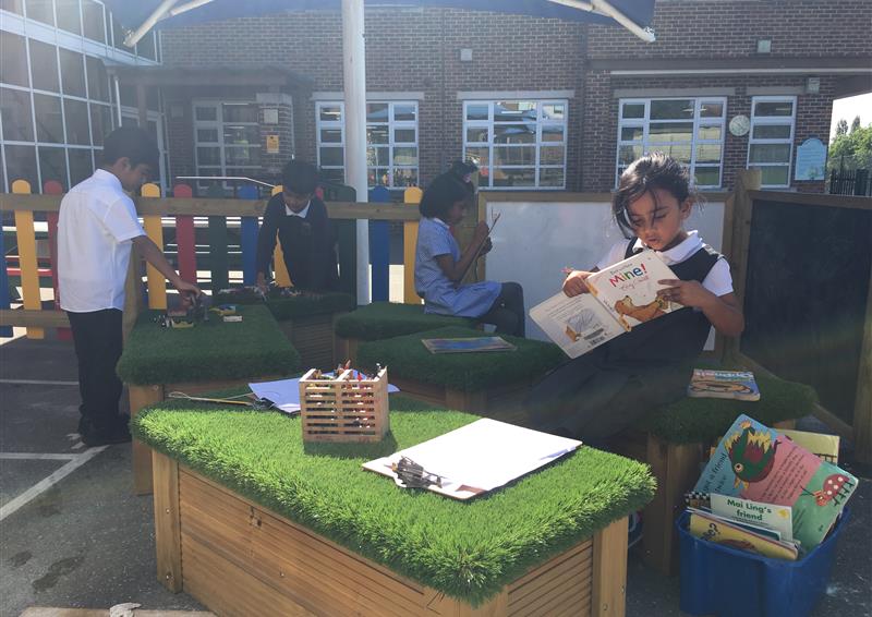 4 children are playing in a seating area with a variety of different materials. The child at the front is reading a book whilst the children at the back are playing with other things. A Large Moveable Artificial Grass-Topped Seat can be seen at the front of the picture.