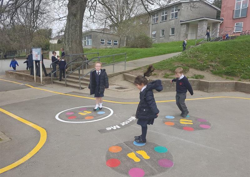 3 children are playing on thermoplastic playground markings. The markings are in the design of follow the leader, with 6 different coloured dots and a pair of footprints in each marking.