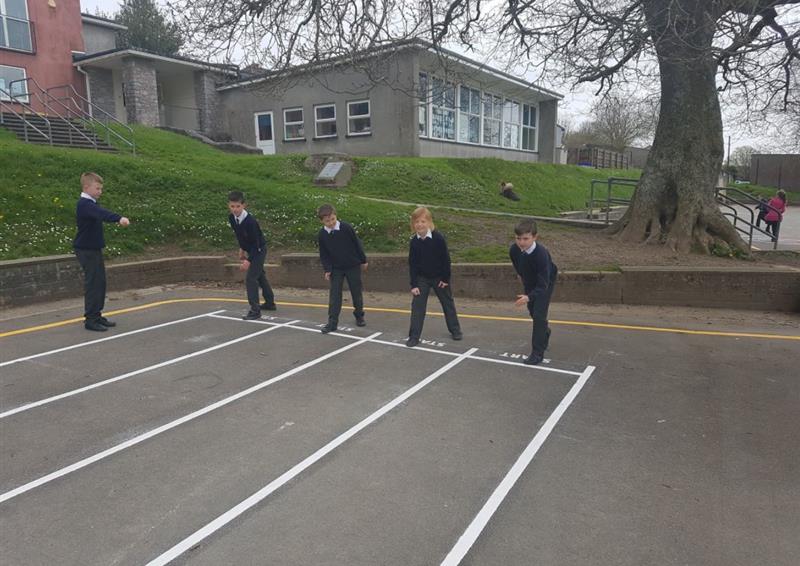 A group of kids are lined up at a 100m race track, which has been marked out by thermoplastic white lines on tarmac. 1 child is stood on the outside of the race, counting the racers down.
