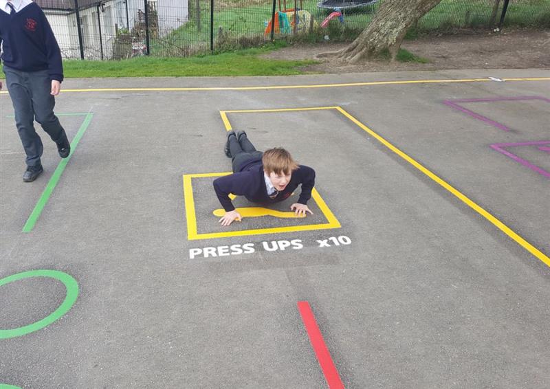 A child has laid down in a yellow thermoplastic box, getting ready to do press ups. A line of white text says "press ups x10". A yellow line is connected to the yellow box, with a red line on the other side.