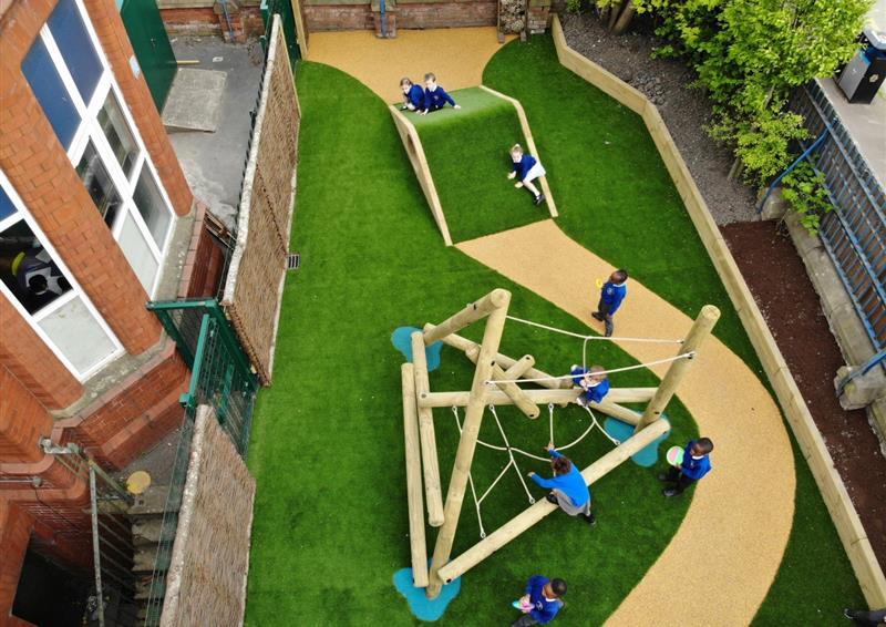 School playground that has a surface of artificial grass, with a wetpour path going through the middle. An Up and Under block has been placed on the path, with a climbing frame placed on the artificial grass.