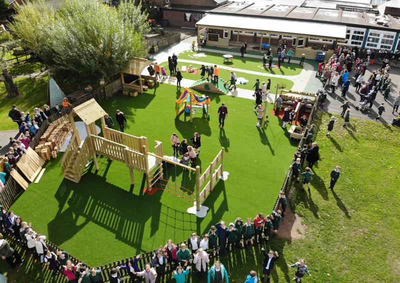 Artificial grass has been used as a surface for a wooden play tower and a variety of other play equipment. A small group of children are playing with the equipment as a large group of children and teachers watch from the wooden fence that surrounds the area.