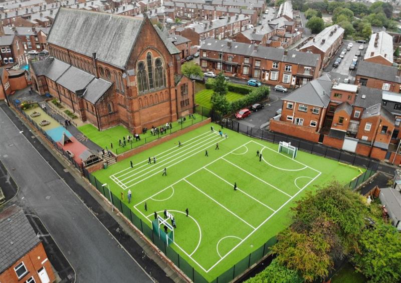 A birds-eye photo of an artificial grass MUGA. There are line markings on the pitch, indicating the MUGA is being used for sports. A group of children are playing on the surface next to the school.