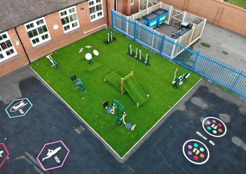 Astroturf installed in the corner of a playground, with gym equipment being placed on the surface, as well as two other pieces of wooden play equipment. Surrounding the artificial grass is a tarmac playground, with thermoplastic play markings.