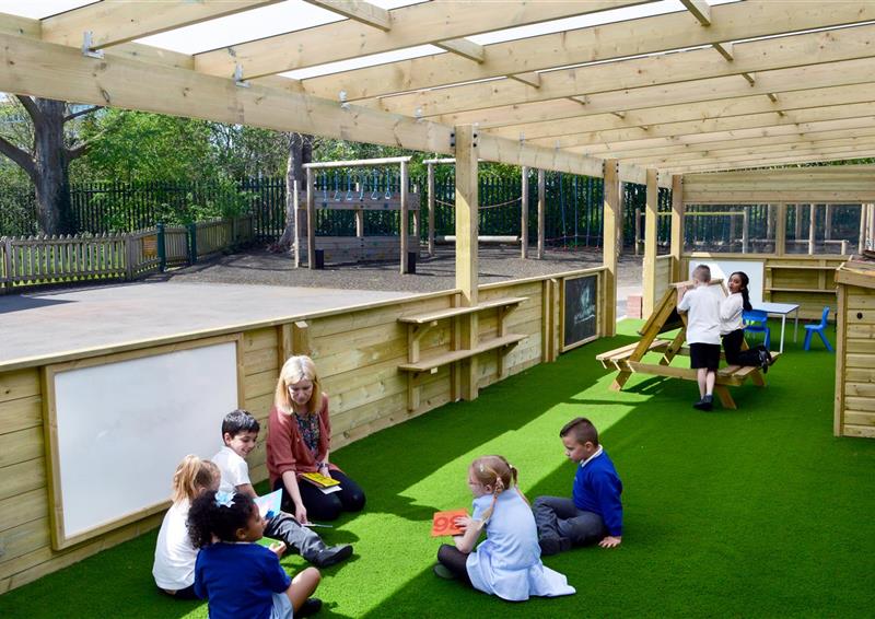 artificial grass installed underneath a wooden canopy, with 5 children sat down around a teacher. 2 children can be seen at the back at a EYFS picnic table.