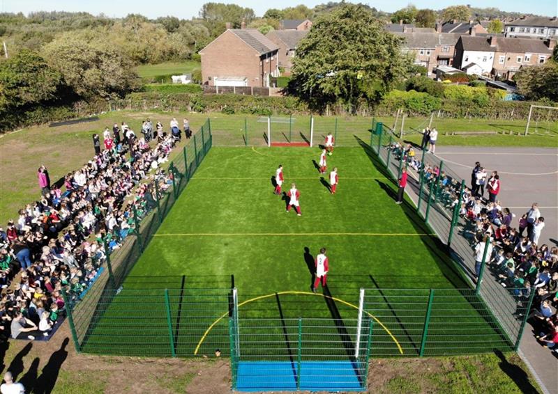 astroturf used as a surface for a MUGA. A group of children are playing football on the MUGA, with hundreds of people watching through the fence.